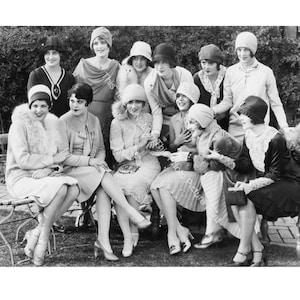 May include: A group of women in 1920s fashion pose for a photo. They are wearing flapper dresses, cloche hats, and fur stoles. The women are smiling and looking at the camera.