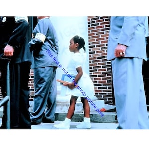 May include: A young girl in a white dress and shoes, holding books, stands between several adults in suits and a police officer. The scene is set in front of a brick building.