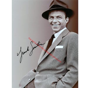May include: Black and white photograph of a smiling man wearing a fedora hat, a patterned suit jacket, and a dark tie. The image includes a signature in black ink. The man is leaning against a neutral-colored background.