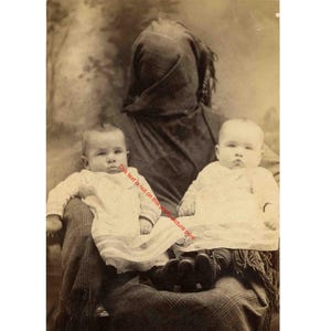 May include: Vintage sepia-toned photograph of a person holding two babies. The babies are wearing white dresses. The person is wearing a dark-colored garment and a head covering. The image has a soft focus and a historical feel.