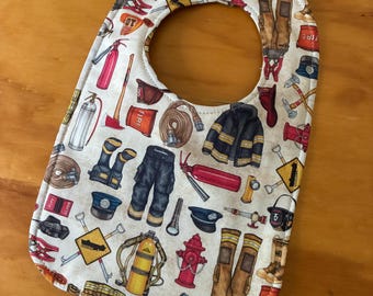 May include: A cream-colored baby bib featuring a repeating pattern of firefighting equipment, including red fire extinguishers, yellow oxygen tanks, black boots, dark trousers, fire hoses, hydrants, helmets, and axes with brown handles.