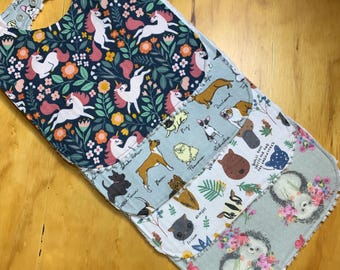 May include: A stack of four baby bibs with different patterns. The top bib features unicorns and flowers on a navy background. The other bibs have dog breeds, floral, and hedgehog designs. The bibs are made of soft fabric.