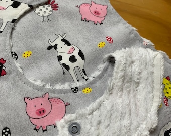 May include: A gray baby bib with a white fleece lining. The bib features a repeating pattern of farm animals, including cows, pigs, and chickens. The bib has two snaps on the shoulder straps.