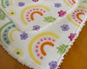 May include: Two white, square baby burp cloths with a colorful rainbow and floral pattern. The rainbows are yellow, orange, and pink. The cloths have a fringed edge and are decorated with purple and pink flowers and green leaves.