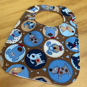 May include: A brown, blue, and white baby bib with a dog pattern. The bib has a round neck opening and is made of soft fabric.