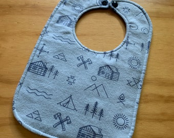 May include: A light blue baby bib with a pattern of camping-themed illustrations, including cabins, mountains, axes, and a compass. The bib has a snap closure.