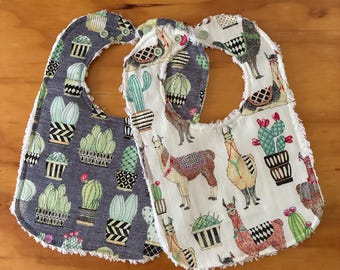 May include: Two baby bibs with snap closures. One bib features a dark gray background with a repeating pattern of various green cacti in pots. The other bib has a white background with a repeating pattern of llamas and cacti in pots.