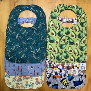 May include: Set of four multi-colored fabric baby bibs with different patterns. The top bib is green with a leaf pattern, the second bib is blue with a fishing lure pattern, the third bib is yellow with a bicycle pattern, and the bottom bib is gray with a cartoon character pattern.