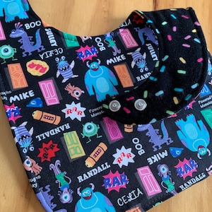 May include: A black bib with a Monsters Inc. print featuring Mike, Sulley, Boo, and Randall. The bib has two silver snaps and a black fleece lining.