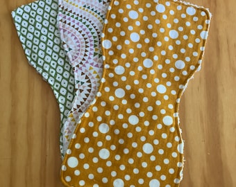 May include: Three cloth diaper inserts with different patterns. One is mustard yellow with white polka dots, another has a geometric design in olive green and white, and the third has a pink, white, and gold triangle pattern.