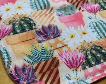 May include: Close-up of a fabric with a repeating pattern of cacti, succulents, and flowers in pots. The design features various shades of green, pink, and white on a beige background with brown stripes and pink polka dots.