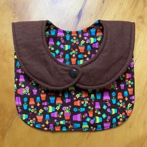 May include: A baby bib with a brown collar and a colorful floral pattern. The bib features a dark brown collar with a single black button closure. The interior fabric has a repeating pattern of potted flowers in various colors.