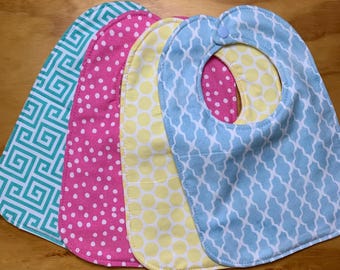 May include: Four baby bibs in various patterns and colors. The bibs include a teal geometric pattern, pink polka dots, yellow polka dots, and a light blue trellis design. Each bib has a curved shape and a neck opening.