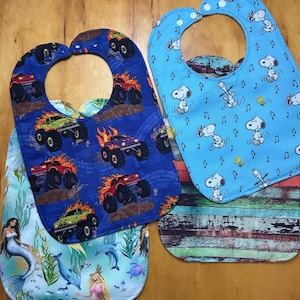 May include: Four baby bibs with different patterns. Two bibs feature a blue background with monster trucks, flames, and large tires. Another bib has a light blue background with Snoopy and musical notes. The fourth bib has a mermaid and ocean theme.
