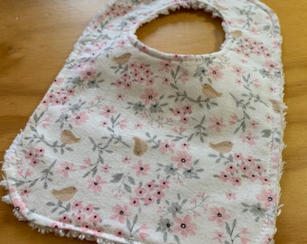 May include: A white baby bib with a pink and gray floral pattern and small brown birds. The bib has a frayed edge.