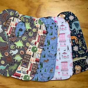 May include: Five different baby bibs with various patterns. The bibs are made of soft fabric and have a unique design. The patterns include dinosaurs, fire trucks, a forest map, a city scene, and a jungle scene.