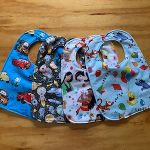 May include: Four baby bibs with different patterns. One is blue with Cars characters, another has a space theme, a third features Disney princesses, and the last is light blue with Winnie the Pooh and balloons.