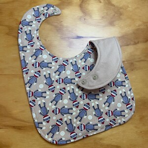 May include: A beige baby bib with a pattern of blue sock monkeys and white buttons. The bib has a brown suede-like collar with two snap closures.