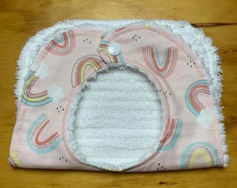 May include: A baby bib with a pink background and a pattern of colorful rainbows and clouds. The bib has a white terry cloth lining and a white fluffy trim. A white button secures the bib.