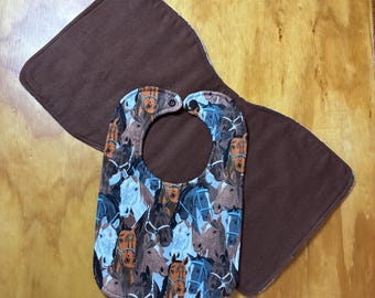May include: A baby bib and burp cloth set. The bib features a pattern of horse faces in various colors, including brown, white, and blue. The burp cloth is a solid brown color. The set is designed for infants and young children.