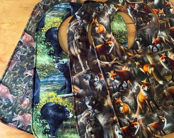 May include: Three fabric baby bibs featuring detailed wildlife prints. The bibs display various animals including wolves, foxes, and bears in forest settings. Each bib has a rounded shape with a neck opening and a small metal snap closure at the top.
