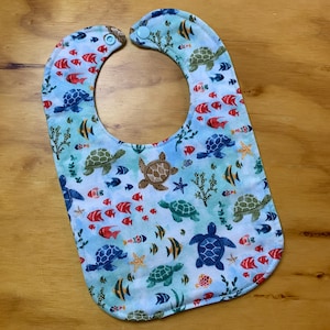 May include: A blue baby bib with a sea turtle and fish pattern. The bib has a blue and white watercolor background with a variety of sea creatures.