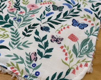 May include: A white cotton bib with a floral pattern featuring butterflies, flowers, and leaves. The bib has a frayed edge.