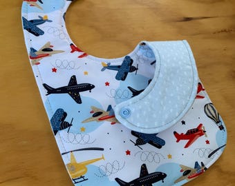 May include: A blue and white baby bib with a pattern of airplanes, stars, and clouds. The bib has a white polka dot trim and a snap closure.