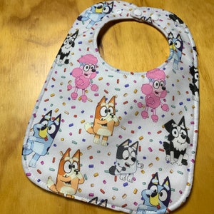 May include: White baby bib with a colorful pattern of cartoon dogs and pink poodles. The bib has a rounded shape with a neck opening and snap closure. The design includes confetti-like shapes.