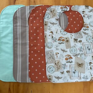 May include: A stack of baby bibs in various patterns and colors. One is solid light blue, another has vertical brown and white stripes, a third is rust-colored with white crosses, and the last features a vintage botanical print with mushrooms and butterflies.