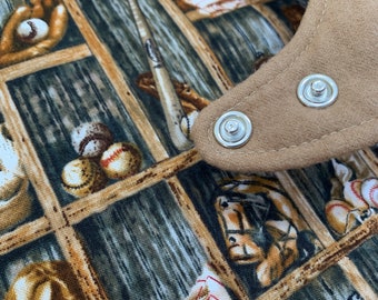 May include: A brown fabric bib with a baseball theme. The bib has a brown felt trim with two silver snaps. The fabric features a repeating pattern of baseball bats, gloves, and balls in a wooden locker room setting.