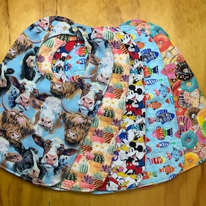 May include: Five colorful fabric bibs with different patterns. One bib features a cow pattern, another has a cactus pattern, another has a Mickey Mouse pattern, another has a fish pattern, and the last one has a donut pattern.
