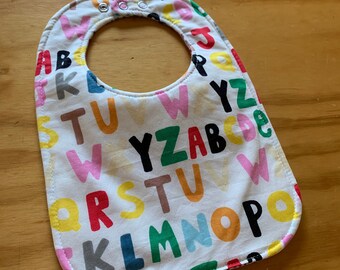 May include: White baby bib with colorful letters of the alphabet printed on it. The bib has a white background with colorful letters of the alphabet printed on it. The letters are in a variety of colors, including red, blue, green, yellow, and orange. The bib has a white background with colorful letters of the alphabet printed on it. The letters are in a variety of colors, including red, blue, green, yellow, and orange.