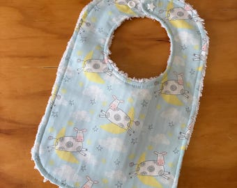 May include: A light blue baby bib featuring a repeating pattern of white cows with pink snouts, yellow crescent moons, white clouds, and small stars. The bib has a white fringed edge and a white plastic snap closure at the top neck opening.