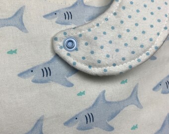 May include: A white baby bib with a blue polka dot trim and a blue shark pattern. The bib has a snap closure on the back.