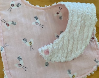 May include: A baby bib with a pink background and a pattern of white birds with black and yellow details. The bib has a white, textured, absorbent section and a pink snap closure.