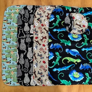 May include: Four different fabric bibs with various patterns. The first bib is light blue with a duck pattern. The second bib is black with a white cat pattern. The third bib is beige with a dog and bone pattern. The fourth bib is black with a green and blue lizard pattern.