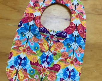 May include: A baby bib with a colorful butterfly and floral pattern. The bib features a white background with a repeating design of butterflies in shades of blue, purple, yellow, orange, and pink. It has a snap closure at the neck.