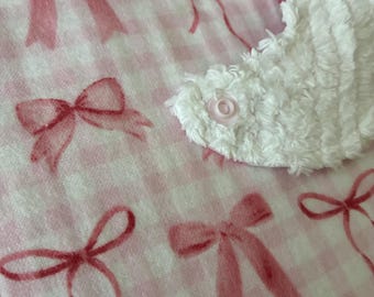 May include: A baby bib featuring a light pink and white gingham pattern with a repeating print of soft pink bows. The bib has a white, textured, terry cloth-like fabric section on the side, secured with a small, round, light pink plastic snap fastener.