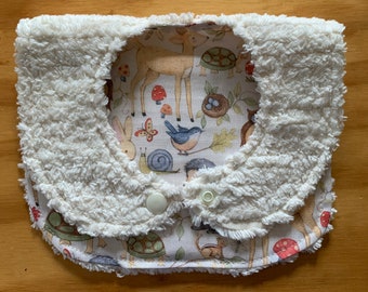 May include: A baby bib with a cream-colored, fluffy border and a woodland animal print interior. The print features deer, turtles, birds, mushrooms, and other forest creatures. The bib has two snap closures.