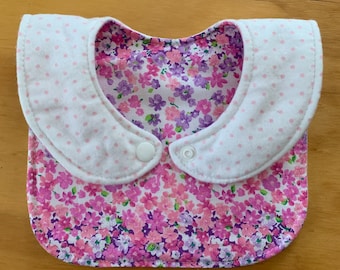 May include: A baby bib with a floral pattern in shades of pink and purple. The bib has a white collar with pink polka dots and two snap closures. The bib is designed to catch spills and protect clothing.