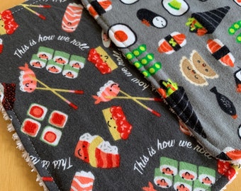 May include: A gray fabric kitchen towel with a sushi theme. The towel has a white border and features a repeating pattern of sushi rolls, chopsticks, and the text "This is how we roll".