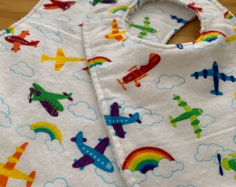 May include: White baby bib and burp cloth set with a colorful airplane and rainbow pattern. The airplanes are in various colors, including red, yellow, green, and blue, with white clouds in the background. The bib has a snap closure.