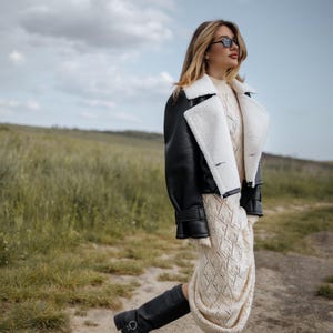 May include: A woman wearing a black leather jacket with white shearling trim, a cream-colored knit dress, and black boots. The outfit is styled with sunglasses. The background is a field with green grass and a cloudy sky.