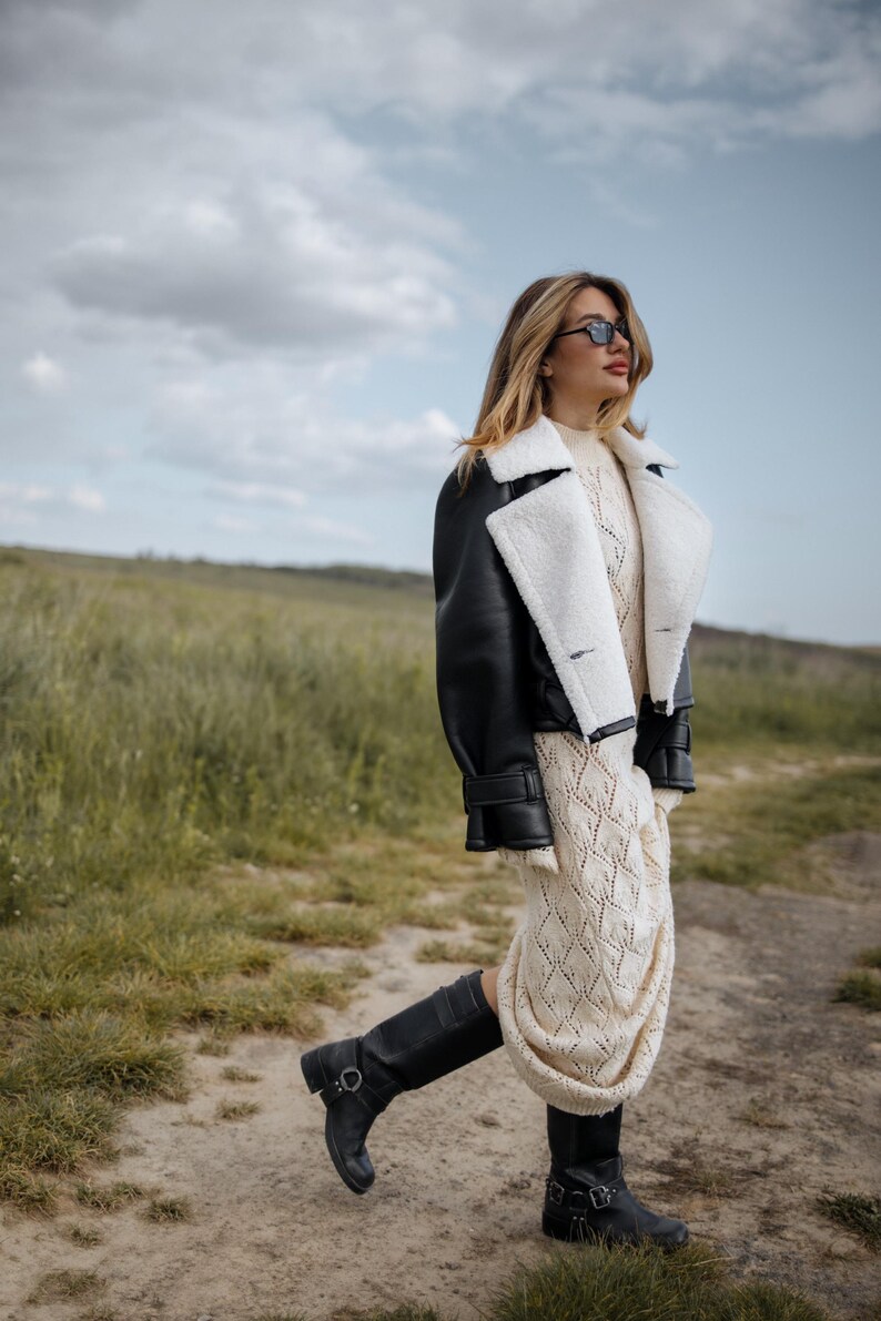 May include: A woman wearing a black leather jacket with white shearling trim, a cream-colored knit dress, and black boots. The outfit is styled with sunglasses. The background is a field with green grass and a cloudy sky.
