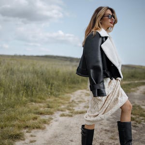 May include: A black leather jacket with a white shearling lining is worn over a cream-colored dress. The outfit is completed with black knee-high boots. The background features a dirt path and a field of tall grass under a cloudy sky.