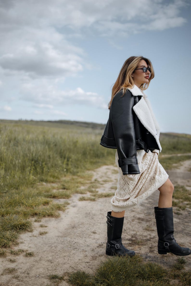 May include: A black leather jacket with a white shearling lining is worn over a cream-colored dress. The outfit is completed with black knee-high boots. The background features a dirt path and a field of tall grass under a cloudy sky.
