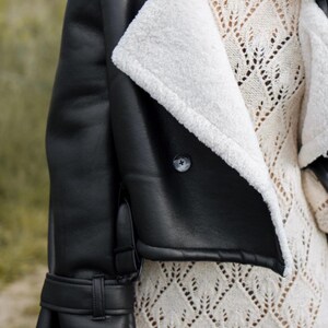 May include: Black faux leather jacket with a white shearling lining and collar. The jacket features a button closure and a buckle detail on the sleeve. The model is wearing a cream-colored knit sweater underneath.
