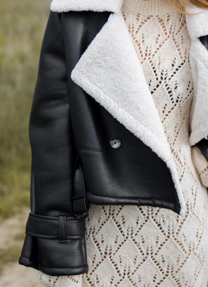 May include: Black faux leather jacket with a white shearling lining and collar. The jacket features a button closure and a buckle detail on the sleeve. The model is wearing a cream-colored knit sweater underneath.