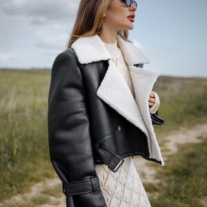 May include: A black leather jacket with a white shearling collar and lining. The jacket has a cropped design with a buckle closure at the waist and cuffs. The model is wearing sunglasses and a cream-colored sweater and dress.